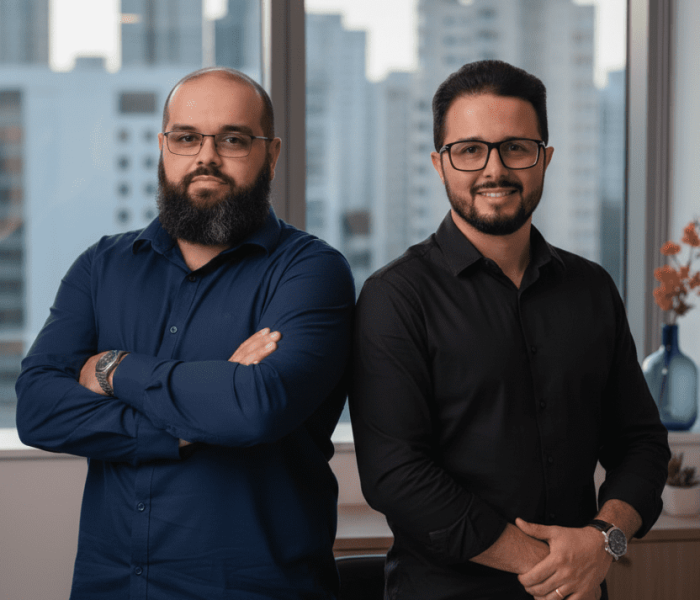 Dois homens profissionais em escritório com vista urbana ao fundo.